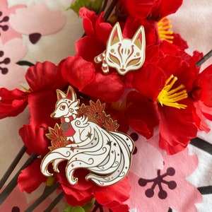 May include: Two gold-tone enamel pins featuring a white fox with a red scarf and a white fox mask. The pins are displayed on a background of red and white fabric flowers.