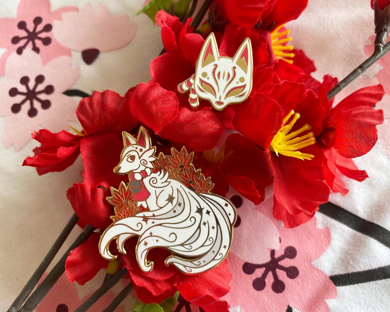 May include: Two gold-tone enamel pins featuring a white fox with a red scarf and a white fox mask. The pins are displayed on a background of red and white fabric flowers.