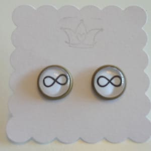 Op de afbeelding: Een paar oorbellen met oneindigheidssymbool bedels. De bedels zijn zilverkleurig met een zwart oneindigheidssymbool op een witte achtergrond. De oorbellen liggen op een witte kaart met een kroonontwerp.