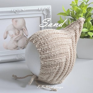 Könnte beinhalten: Eine gestrickte Kapuze in Sandfarbe mit spitzer Oberseite und Bindebändern. Die Kapuze wird auf einer weißen Form präsentiert. Das Wort "Sand" ist kursiv auf dem Bild geschrieben. Der Text "PaBoWool Design" ist ebenfalls sichtbar.