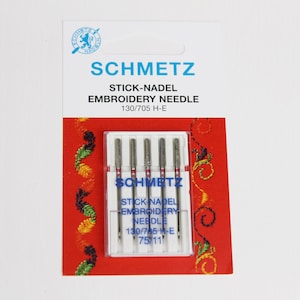 Könnte beinhalten: Eine Packung mit fünf silbernen Sticknadeln in einem durchsichtigen Kunststoffbehälter. Die Verpackung ist mit "SCHMETZ STICK-NADEL EMBROIDERY NEEDLE 130/705 H-E" beschriftet.