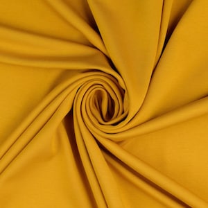 May include: A close-up of a smooth, mustard yellow fabric. The fabric is folded and draped in a way that creates a swirling pattern.