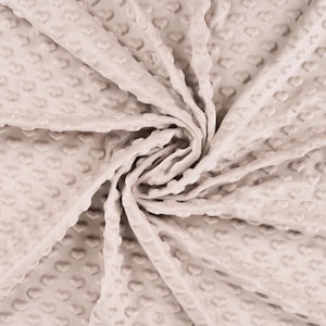Könnte beinhalten: Nahaufnahme eines beigefarbenen Stoffes mit einem strukturierten Muster aus kleinen Herzen. Der Stoff ist weich und plüschig und eignet sich perfekt für eine Babydecke oder eine Tagesdecke.