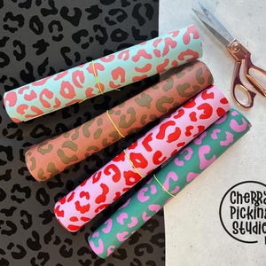 May include: Four rolled patterned fabrics in pink, red, green, and brown with a leopard print design. A pair of rose gold scissors rests on the right. The fabrics are secured with yellow rubber bands. A black surface with a leopard print pattern is on the left.
