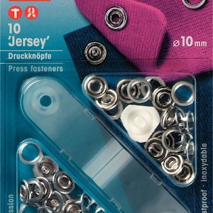 Könnte beinhalten: Eine Packung mit 10 silberfarbenen, 10 mm großen, rostfreien Jersey-Druckknöpfen. Die Packung enthält eine transparente Plastikfolie mit den Druckknöpfen und ein weißes Plastikteil. Die Packung ist mit "10 Jersey' Druckknöpfe Press fasteners" und "rostfrei rustproof inoxydable" beschriftet.