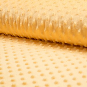 Könnte beinhalten: Nahaufnahme eines gelben Minky-Stoffes mit einem strukturierten Punktmuster. Der Stoff ist weich und plüschig, perfekt für Babydecken, Quilts und andere Nähprojekte.