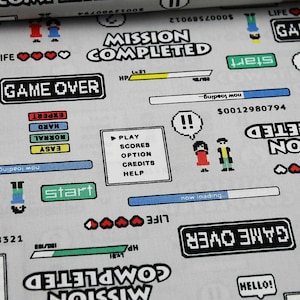 Könnte beinhalten: Ein weißer Stoff mit einem sich wiederholenden Muster aus pixelartigen Videospielgrafiken. Die Grafiken beinhalten die Wörter "Mission abgeschlossen", "Game Over", "Start", "Leben", "Hilfe", "Credits", "Option", "Punkte", "Spielen" und "Lädt".