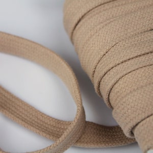 May include: A spool of tan-colored, flat cotton tape. The tape is tightly wound in a roll, with a portion unraveled, showing its woven texture. The neutral color and flat design suggest its use in various crafting or sewing projects.