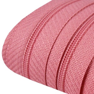 Könnte beinhalten: Nahaufnahme einer Rolle rosa Reißverschlussband. Die Reißverschlusszähne sind am Rand des Bandes sichtbar, und der Stoff hat eine gewebte Textur. Die Farbe ist ein gedämpftes, staubiges Rosa.