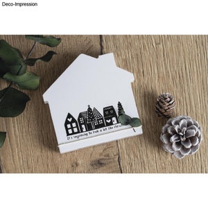 Könnte beinhalten: Eine weiße, hausförmige Holzdekoration mit einem schwarz-weißen Dorfdesign am unteren Rand. Der Text "It's beginning to look a lot like christmas" ist darunter gedruckt. Zwei Tannenzapfen und Grünzeug liegen daneben.