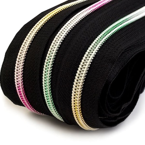 May include: Close-up of four black zipper tapes with different coloured metal teeth. The teeth are gold, silver, green, and pink.