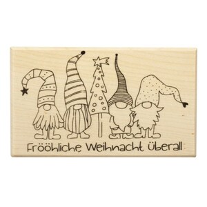 May include: A wooden stamp featuring a design of four gnomes wearing hats and standing in front of a Christmas tree. The text "Fröhliche Weihnachten überall" is printed below the gnomes.