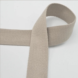 Puede incluir: Una cinta de algodón beige, de aproximadamente 2,5 cm de ancho, se muestra doblada en forma de ocho. La cinta está tejida con un patrón texturizado.