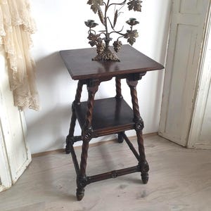 May include: A dark brown, antique wooden side table with a lower shelf and intricately carved legs. A decorative brass candelabra with leaf and grape details sits on top. The candelabra has multiple candle holders.