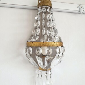 Puede incluir: Aplique de pared antiguo con marco de metal dorado y detalles de cristal transparente. El aplique presenta una corona decorativa en la parte superior y colgantes de cristal. La l&aacute;mpara est&aacute; montada en una pared blanca.