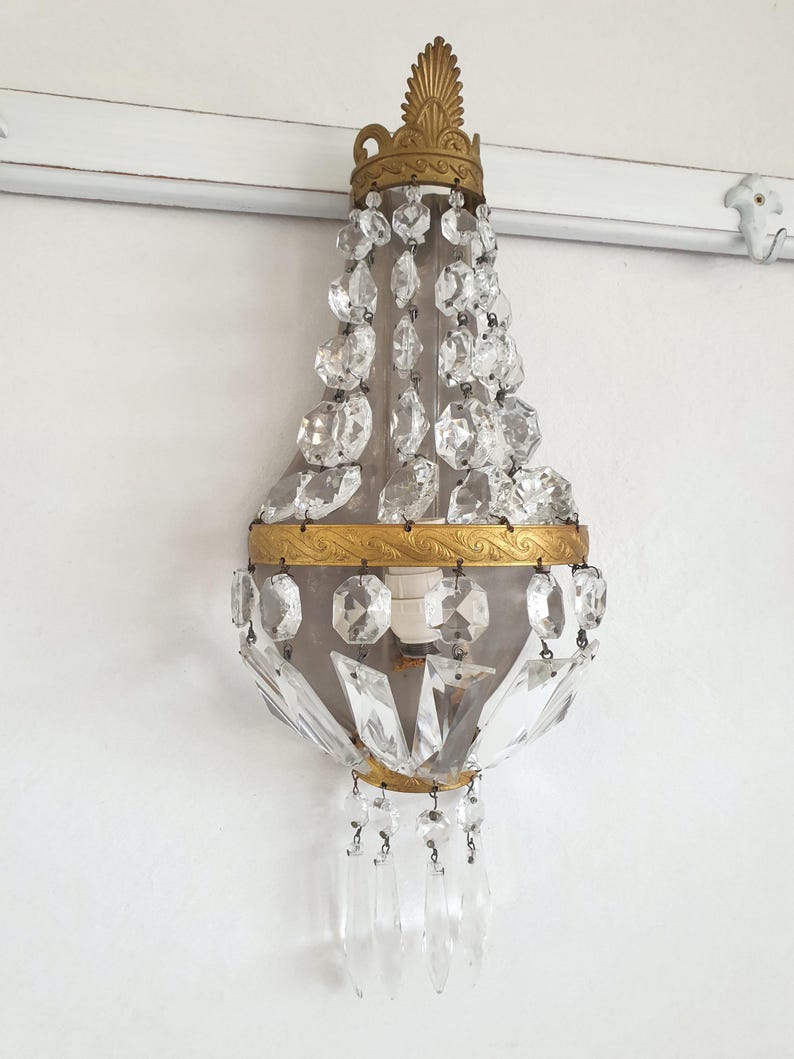 Puede incluir: Aplique de pared antiguo con marco de metal dorado y detalles de cristal transparente. El aplique presenta una corona decorativa en la parte superior y colgantes de cristal. La l&aacute;mpara est&aacute; montada en una pared blanca.