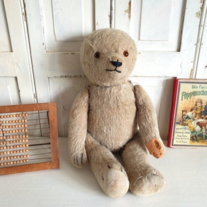 May include: A vintage, beige teddy bear with black eyes and a black nose sits on a white surface. A wooden abacus and a red and yellow book titled "Die Fleissige PuppenSchneiderin" are also on the surface.