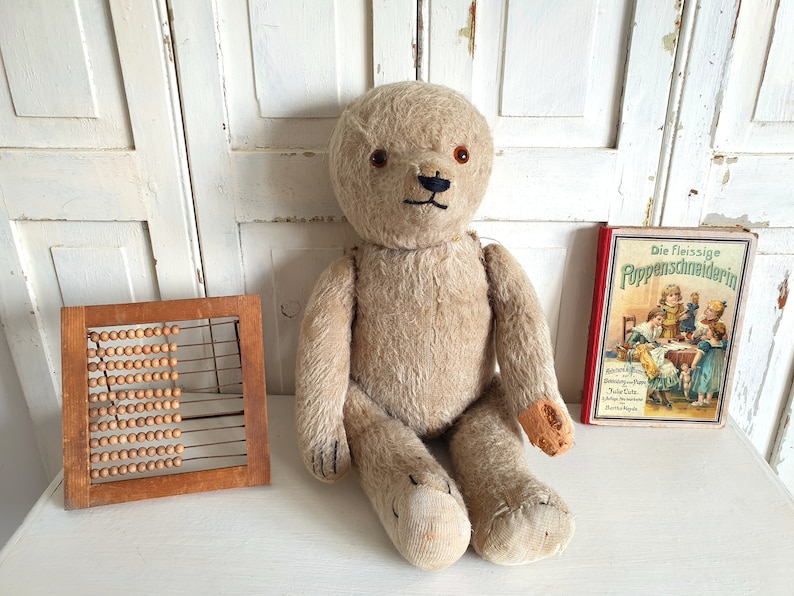 May include: A vintage, beige teddy bear with black eyes and a black nose sits on a white surface. A wooden abacus and a red and yellow book titled "Die Fleissige PuppenSchneiderin" are also on the surface.
