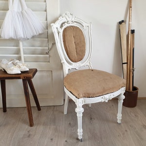 Puede incluir: Una silla antigua pintada de blanco con un asiento de tela marr&oacute;n y un respaldo tallado. La silla est&aacute; sobre un suelo de madera.