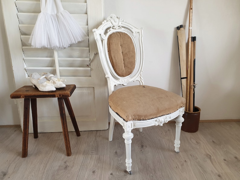 Puede incluir: Una silla antigua pintada de blanco con un asiento de tela marr&oacute;n y un respaldo tallado. La silla est&aacute; sobre un suelo de madera.