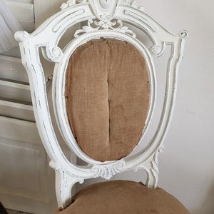 Puede incluir: Silla blanca pintada con un marco de madera tallada y un asiento de tela marr&oacute;n. La silla tiene un acabado desgastado y un aspecto usado.