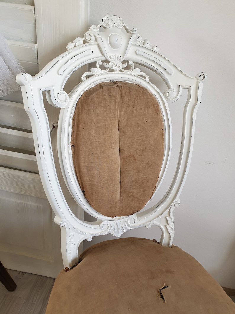 Puede incluir: Silla blanca pintada con un marco de madera tallada y un asiento de tela marr&oacute;n. La silla tiene un acabado desgastado y un aspecto usado.