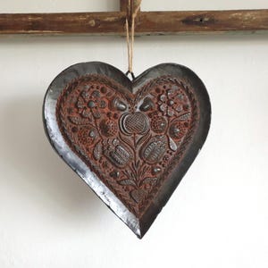 May include: A heart-shaped, rustic metal wall hanging. The dark gray metal frame surrounds a detailed, embossed design in shades of brown and rust. The embossed design features floral and botanical elements. A natural twine loop is attached for hanging.