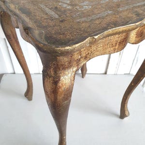 May include: A small, antique side table with a square top and curved legs. The table is painted in a distressed gold finish, with visible wear and tear adding to its vintage charm. The table's dimensions are approximately 20 inches in height and 18 inches wide.