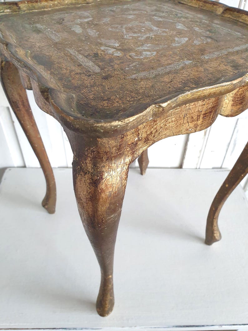 May include: A small, antique side table with a square top and curved legs. The table is painted in a distressed gold finish, with visible wear and tear adding to its vintage charm. The table's dimensions are approximately 20 inches in height and 18 inches wide.