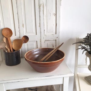 Könnte beinhalten: Eine rustikale Küchenszene mit einer großen braunen Keramikschüssel und Holzlöffeln. Eine Sammlung von Holzlöffeln befindet sich in einem dunklen Keramiktopf. Eine dekorative Vase mit getrocknetem Lavendel ist ebenfalls vorhanden.