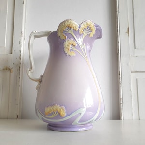 Könnte beinhalten: Eine lavendelfarbene Keramikkrug mit gerüschtem Rand und gebogenem Griff. Der Krug ist mit gelben und weißen floralen Designs verziert. Der Krug ist ein dekorativer Gegenstand.