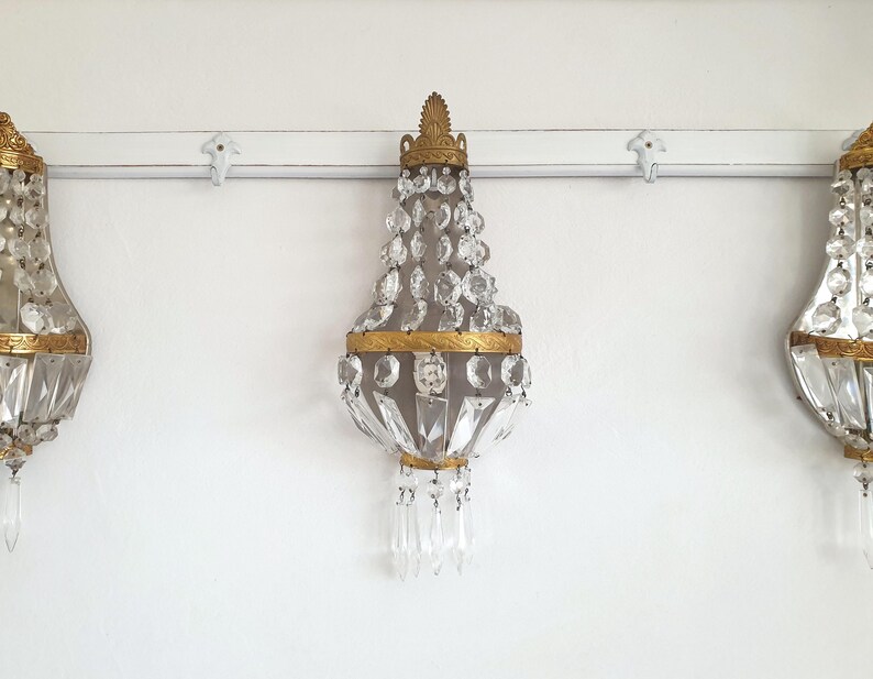 Puede incluir: Tres apliques de pared ornamentados con detalles de cristal y dorado. Los apliques tienen forma de l&aacute;grima, con cristales transparentes que caen de una corona dorada. Est&aacute;n montados en una pared blanca.
