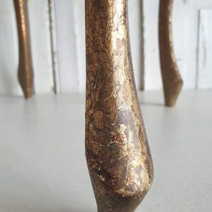 May include: Close-up of a furniture leg with a distressed gold leaf finish. The leg is curved and tapers towards the bottom, showcasing a vintage or antique aesthetic. The gold leaf has a textured, aged appearance.