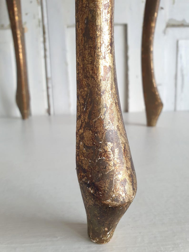 May include: Close-up of a furniture leg with a distressed gold leaf finish. The leg is curved and tapers towards the bottom, showcasing a vintage or antique aesthetic. The gold leaf has a textured, aged appearance.