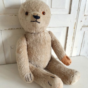May include: A vintage, beige teddy bear with brown button eyes and a black stitched nose. The bear is sitting on a white surface and has some wear and tear, particularly on one paw.