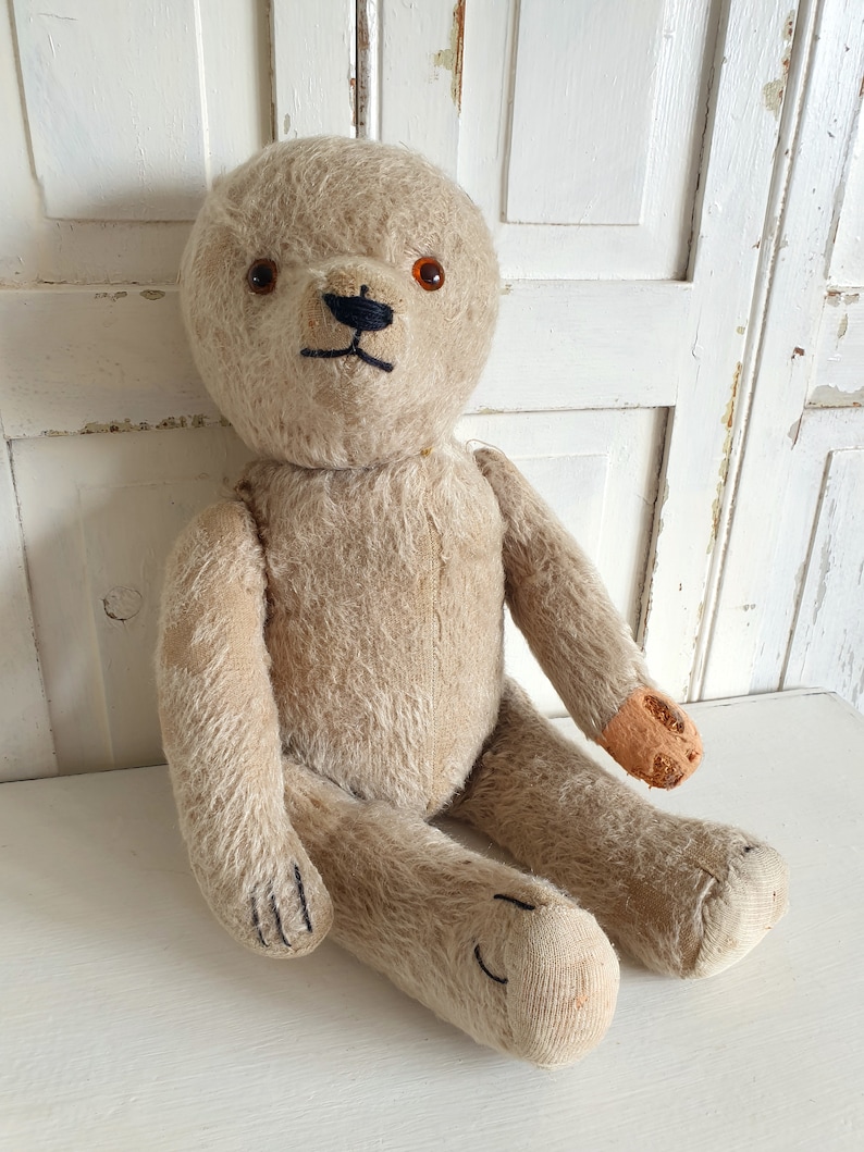 May include: A vintage, beige teddy bear with brown button eyes and a black stitched nose. The bear is sitting on a white surface and has some wear and tear, particularly on one paw.