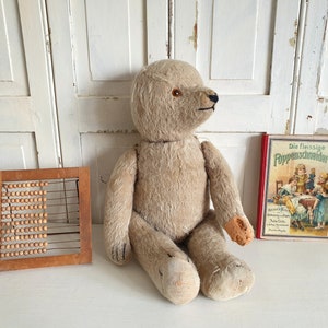 May include: A vintage, beige teddy bear with button eyes sits on a white surface next to a wooden abacus and a book titled "Die Fleissige PuppenSchneiderin".