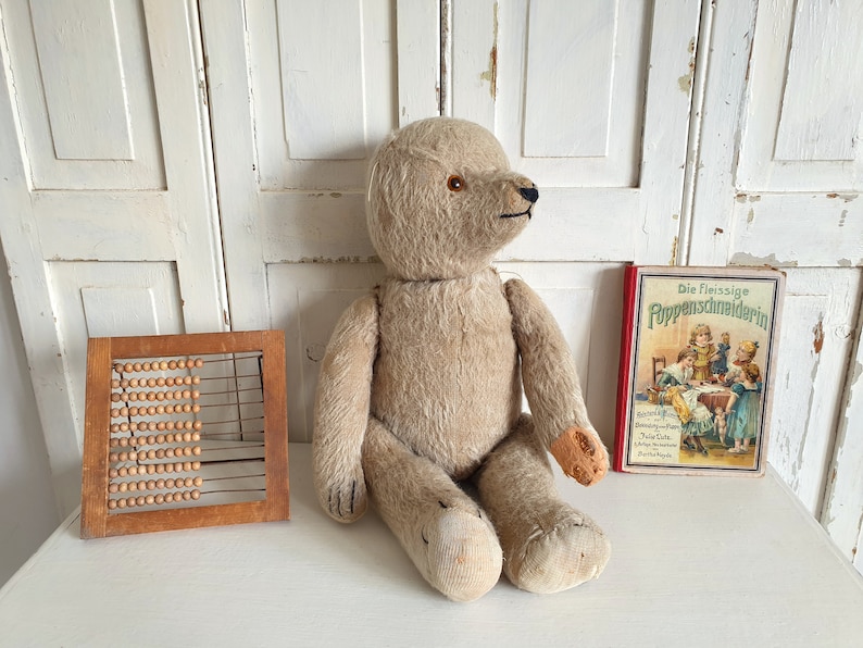 May include: A vintage, beige teddy bear with button eyes sits on a white surface next to a wooden abacus and a book titled "Die Fleissige PuppenSchneiderin".