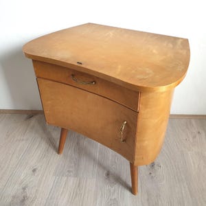 Op de afbeelding: Een licht houten nachtkastje met een gebogen bovenkant en één lade. De kast heeft messing handgrepen en staat op vier poten.