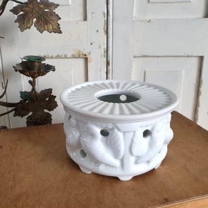 Può includere: Un scaldino per tè in ceramica bianca con la parte superiore rotonda e scanalata e decorazioni a foglie e frutta. Lo scaldino ha diversi piccoli fori sui lati e poggia su una superficie di legno. Un candelabro decorativo in metallo è sullo sfondo.