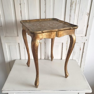May include: A small, square antique table with a gold finish. The table has a detailed top and curved legs. The table is approximately 24 inches tall and 18 inches wide.