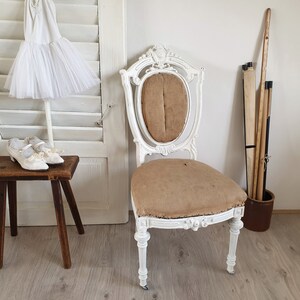 Puede incluir: Una silla antigua pintada de blanco con un asiento de tela marr&oacute;n y detalles tallados. La silla est&aacute; sobre un suelo de madera con una puerta de contraventanas blancas y un peque&ntilde;o taburete de madera en el fondo.