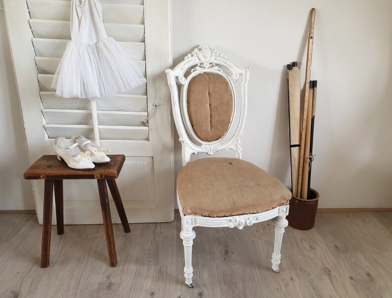 Puede incluir: Una silla antigua pintada de blanco con un asiento de tela marr&oacute;n y detalles tallados. La silla est&aacute; sobre un suelo de madera con una puerta de contraventanas blancas y un peque&ntilde;o taburete de madera en el fondo.