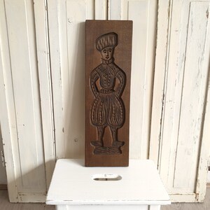 Puede incluir: Un molde de madera rectangular con una imagen tallada de una persona con ropa hist&oacute;rica. La figura lleva un sombrero, una camisa con cuello y pantalones abombados. La madera tiene un tono marr&oacute;n c&aacute;lido.