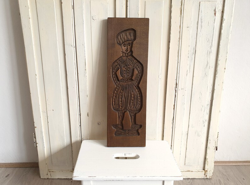 Puede incluir: Un molde de madera rectangular con una imagen tallada de una persona con ropa hist&oacute;rica. La figura lleva un sombrero, una camisa con cuello y pantalones abombados. La madera tiene un tono marr&oacute;n c&aacute;lido.