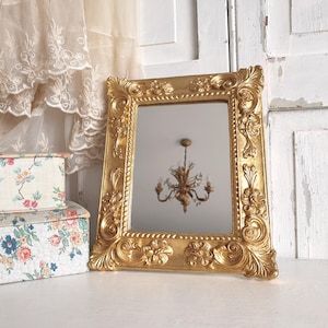 Peut inclure: Un miroir encadré doré et orné de forme rectangulaire. Le cadre présente des motifs floraux et volutes détaillés. Le miroir reflète un lustre. Des boîtes décoratives à motifs floraux sont en arrière-plan.