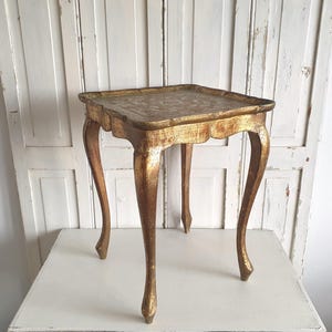 May include: A small, antique gold-painted table with a square top and curved legs. The table has a decorative, ornate design on the tabletop and a distressed finish, giving it a vintage appearance. The table is approximately 24 inches tall.