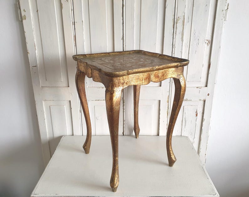 May include: A small, antique gold-painted table with a square top and curved legs. The table has a decorative, ornate design on the tabletop and a distressed finish, giving it a vintage appearance. The table is approximately 24 inches tall.