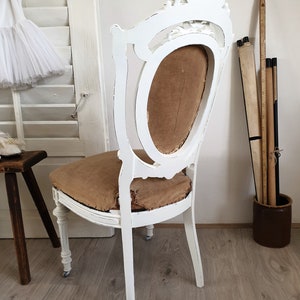 Puede incluir: Silla de madera pintada de blanco con respaldo tallado y asiento de tela marr&oacute;n. La silla est&aacute; en una habitaci&oacute;n con paredes blancas y suelo de madera.