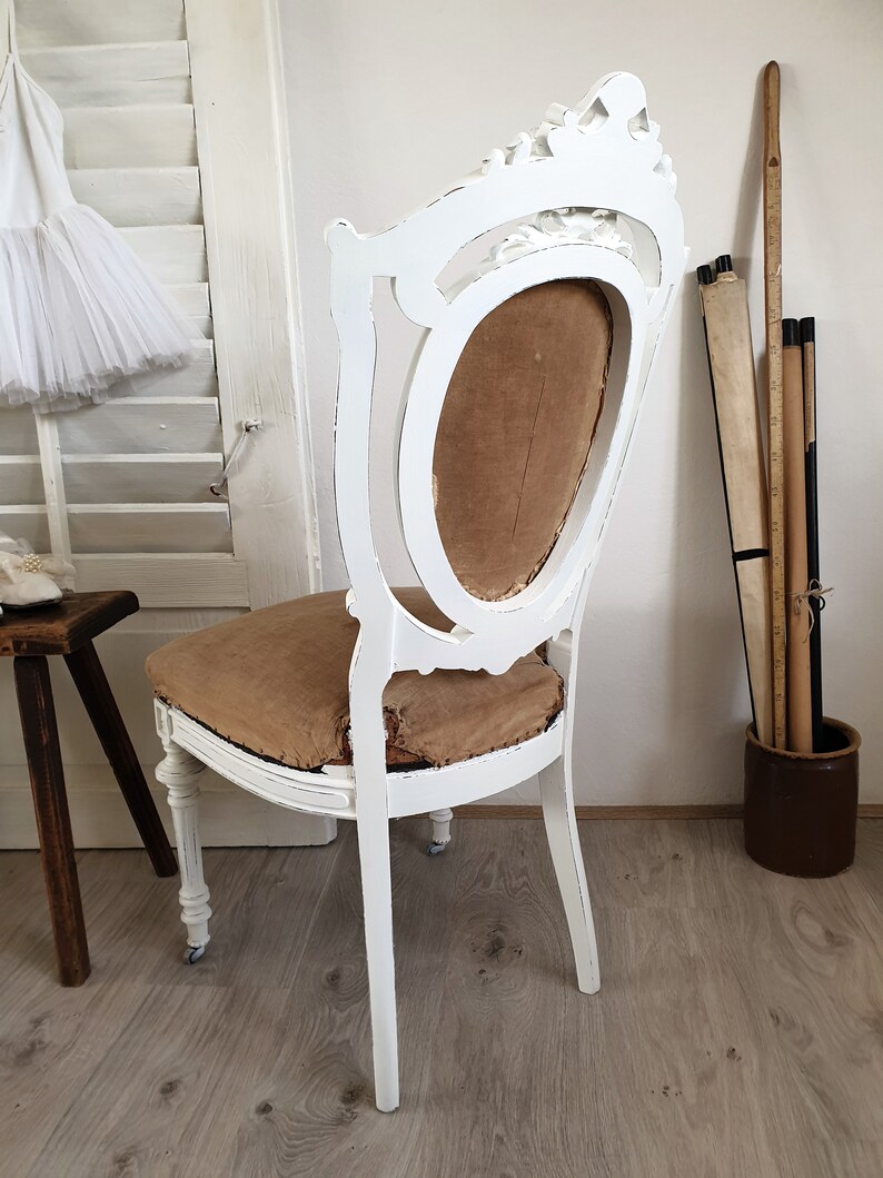 Puede incluir: Silla de madera pintada de blanco con respaldo tallado y asiento de tela marr&oacute;n. La silla est&aacute; en una habitaci&oacute;n con paredes blancas y suelo de madera.